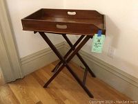 Full view of wooden butler's tray on folding stand against wall on wooden floor.