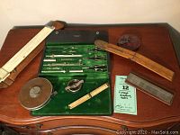 Overview of all drafting and measuring tools on wooden table including case, rulers, tape measure, and surface reference tools.