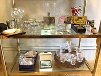 Wide shot of vintage barware displayed on glass shelves showing silver overlay coupes, crystal goblets, flask, glass mugs with red logos, and boxed cocktail tools.