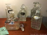 Front and back view of five glass decanters arranged on wooden surface showing various stoppers and liquor tags.