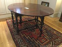 Front view of antique oval dining table with gateleg design, showing the wood tabletop and attached legs standing on a patterned rug.