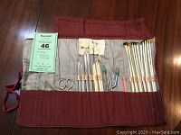 Full view of vintage knitting kit containing knitting needles, cable needles, a pair of scissors, and multiple knitting tool slots in a burgundy fabric roll-up case.