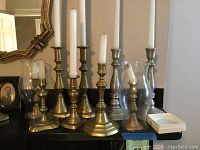 Photo showing ten vintage brass and pewter candlesticks in various sizes on a black mantel, some with white taper candles and glass hurricane chimneys.