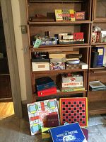 Photo of multiple shelves filled with vintage and newer boxed puzzles and games, some stacked. Also shows large classic game boards for Parcheesi, checkers, and puzzles leaning against shelf.