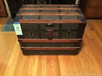 Front view of antique trunk showing black paint, wooden slats, metal latch, and wheels beneath.
