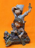 Front and left side view of Elisa Acisclo figurine titled 'Adventure' showing two children, one standing with wooden sword, the other crouching with wooden rifle on raft base against orange background.