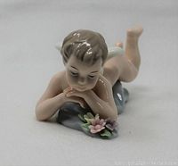 Close-up photo of porcelain angel figurine lying on stomach with head resting on clasped hands, with visible flower details on base.
