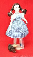 Front view of Dorothy doll wearing blue gingham dress and white blouse, standing with Toto dog figurine on a white display stand. Doll's legs wrapped in protective plastic.