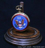 Gold-tone pocket watch showing the Presidential Seal on the back, hanging on the oak stand post without the glass dome cover.