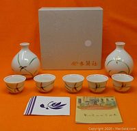 Photo shows entire mini porcelain sake set including two flasks, five small cups, two paper items, and original box on orange background.