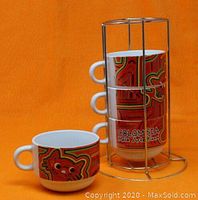 Four ceramic mugs stacked vertically in gold-tone metal wire rack against orange background, showing detailed cat art motifs and 'Colombia Mola Arte Kuna' text.