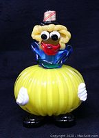 Front view of handcrafted Murano glass clown figurine with yellow ribbed body, blue collar, blonde hair, black top hat, and big red lips smiling face.