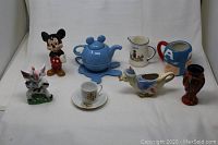 Photo showing entire group of figurines and pottery items, including Mickey Mouse figurine, blue teapot, baby pitcher, Captain America mug, rabbit figurine, bird-shaped creamer, small vase, and cup with saucer.
