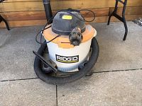 Front view of Rigid shop vacuum showing white body, orange lid, black hose wrapped around base and black power cord.