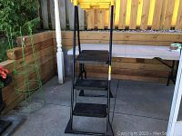 Full view of the folded ladder standing on a patio area, showing its black metal frame and yellow plastic top shelf.