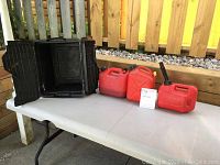 Photo showing three red gas cans and one black utility tote placed on white table outdoors.