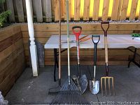 Six yard tools arranged standing on concrete floor against wooden fence and folding table behind them.