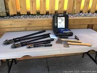 Full lot arranged on white folding table outdoors under daylight, showing tools including jigsaw case, bolt cutters, hatchets, and hammer.