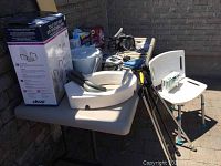 Wide view of medical supplies and home care accessories on a plastic folding table outdoors against brick wall, showing toilet seat riser box, white plastic shower chair, walking canes, CPAP machine, and blood pressure monitor.