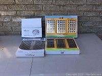 Photo showing the light fixture box and packages of floor register covers on a table against a brick wall.