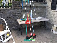 Wide view showing entire lot of cleaning tools arranged around table outside, including vacuum, brooms, dustpan, mops, and duster.
