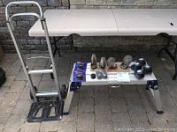 Photo showing lightweight aluminum hand truck dolly, folding step platform, and various casters arranged on paving stones next to a table.