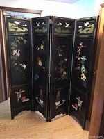 Four-panel black lacquered room divider with colorful birds and flowers painted on the front side, shown angled in room.