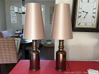 Two modern accent lamps on a table, with ceramic oiled bronze finish bases and beige silk-like tapered shades, unlit, showing overall condition.