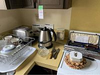 Countertop with Cuisinart toaster, electric kettle, dish rack with dishes and mugs, assorted cutlery, glass ashtray, and decorative kitchen items.