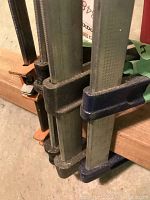 Four metal clamps with steel bars, assembled and resting on wooden beam with varied handle materials including wood and red plastic.