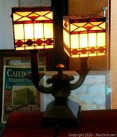 Lamp shown with both stained glass square shades lit, front angle view of bronze base and dual arms.