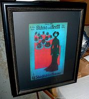 Photo of the full framed poster showing the gothic styled figure and devil heads on plates on a red stand with a blue background and black frame.