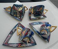 Four triangular plates with abstract multicolor patterns and gold-tone handles paired with four matching cups and bowls.