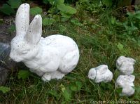 Large white concrete mother rabbit and three smaller baby rabbits on grass with foliage background