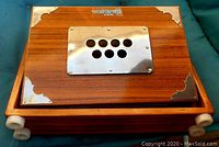 Top view of wooden squeeze box showing metal grille with circular holes and metal corner reinforcements, brand name visible on top.