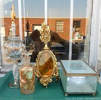Group of four decorative art glass perfume bottles and a mirrored glass jewelry box on a green surface in front of a window, showing details of gold leaf and hand-painted designs.