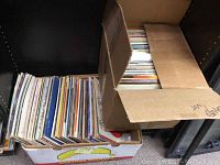 Photo showing two cardboard boxes densely packed with numerous vinyl records placed vertically; the boxes show some wear; the records' covers are facing upwards and outwards, hinting at a varied collection.