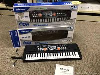 Two BigFun 49-key rechargeable electronic keyboards in boxes, one keyboard out with power cord attached and user manual visible.