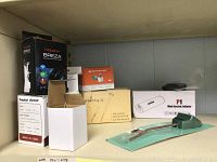 Six new, boxed electronic and home improvement items arranged on a shelf, including aquarium air pump, heat lamp, smart lighting, mini inflator, and solar light.