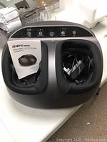 Foot massager placed on table showing top and front control panel with foot compartments and manual.