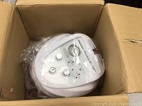 Vacuum therapy machine main unit inside cardboard box, white with control knobs and gauge, wrapped in plastic.