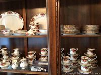 Glass-front cabinet shelf displaying multiple dinner plates, salad plates, cups, saucers and bowls in Old Country Roses pattern