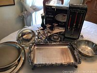 Overview of all silver plated items arranged on table including candleholders, salt and pepper shakers, cake tray, serving platters, and a colander-style bowl.