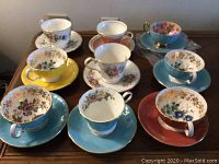 Nine assorted Aynsley bone china teacups and saucers arranged on table, showing variety of exterior colors and interior patterns
