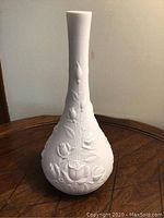 Full view of white Kaiser vase with tall neck and embossed rose pattern on rounded base, placed on wood surface.