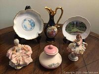 Five collectible items on wood table: two porcelain figurines with fabric accents, two Noritake Nippon plates on stands, small blue and gold Limoges water pitcher, and small jar with lid