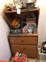 Full view of a wooden cabinet packed with assorted cookbooks, mixing bowls, baking pans, strainers, and kitchen utensils.
