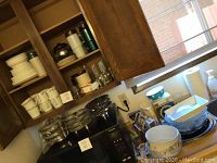Kitchen cabinet with Corelle dishes, mugs, glassware, and pots stacked on stove and counter