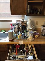 Overall view of kitchen countertop with assorted kitchen items including bowls, thermoses, food processor attachments, and utensils organized on the counter and in a drawer below.