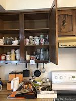 Open wooden kitchen cabinet showing ceramic mugs, glass tumblers, cutting boards, and kitchen utensils on counter.